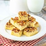 Squares of pecan-topped fudge stacked on a white plate, with a glass and jug of milk in the background on a red checkered cloth.