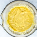 A glass mixing bowl filled with yellow cake batter sits on a white surface.