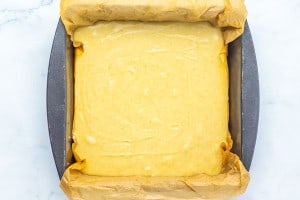 Square baking pan lined with parchment paper, filled with an even layer of uncooked yellow cake batter, on a white surface.