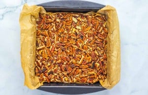 A baking pan lined with parchment paper contains a baked dessert topped with a layer of chopped pecans.