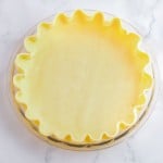 A glass pie dish filled with an unbaked pie crust, edges crimped, on a white marble surface.
