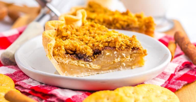 A slice of double-layer pie with a crumb topping sits on a white plate, showing layers of filling and crust, with a red checkered cloth underneath.