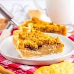 A slice of pie with a crumb topping on a white plate, accompanied by round crackers, with a glass of milk and another pie slice in the background.