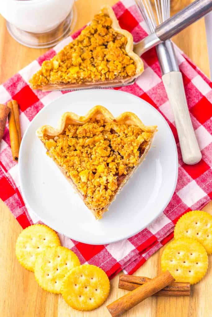 A slice of pie with a crumb topping on a white plate, surrounded by round crackers, cinnamon sticks, utensils, and a red and white checkered cloth.