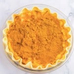 Unbaked pie with a crimped crust in a glass dish, topped with a layer of brown cinnamon powder on a white marble surface.