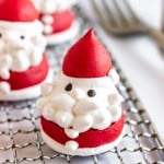 A Santa-shaped meringue cookie with red hat and coat, white beard and trim, and black eyes, sitting on a metal cooling rack.