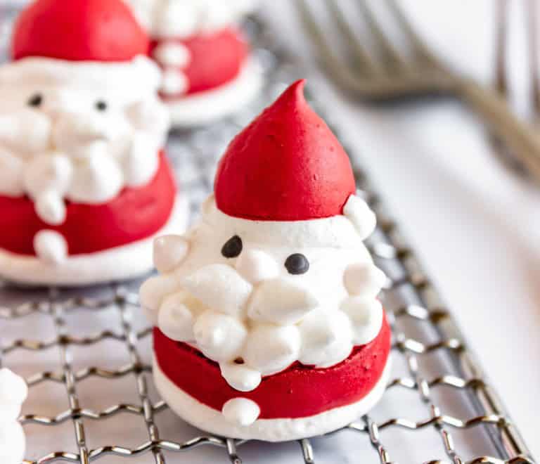 A Santa-shaped meringue cookie with red hat and coat, white beard and trim, and black eyes, sitting on a metal cooling rack.