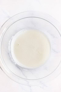 A clear glass bowl filled with a small amount of frothy, white liquid sits on a white marble surface.