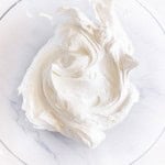 A clear glass bowl containing a large dollop of thick, glossy white whipped cream or frosting on a white marble surface.