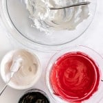 Four bowls of icing in white, red, black, and off-white colors, each with a spoon, are arranged on a white surface.