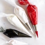 Four piping bags filled with white, red, and black icing, each fitted with metal tips, are arranged on a white marble surface.