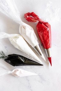 Four piping bags filled with white, red, and black icing, each fitted with metal tips, are arranged on a white marble surface.