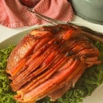 Sliced glazed ham served on a bed of curly green lettuce, with a carving fork and knife nearby and a bowl of cranberry sauce in the background.