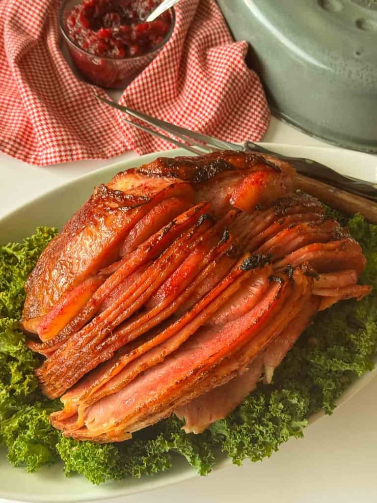 Sliced glazed ham served on a bed of curly green lettuce, with a carving fork and knife nearby and a bowl of cranberry sauce in the background.