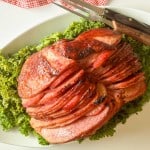 Sliced glazed ham arranged on a bed of leafy greens, served on a white platter with two carving utensils beside it.