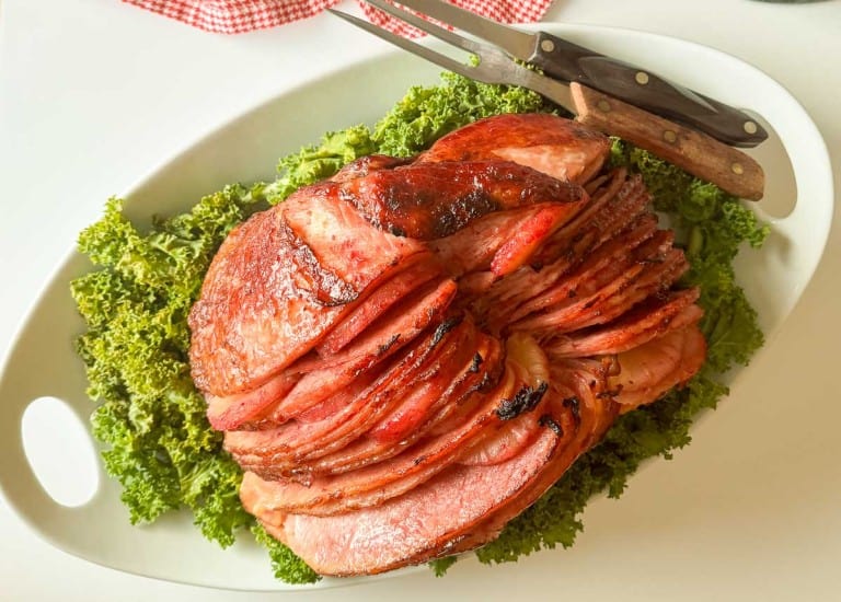 Sliced glazed ham arranged on a bed of leafy greens, served on a white platter with two carving utensils beside it.