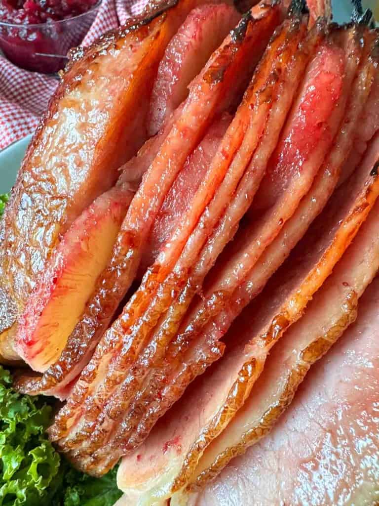 Close-up of sliced glazed ham arranged on a plate, with a side of leafy greens visible in the corner.