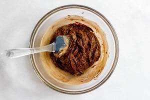 A glass bowl containing thick chocolate batter being mixed with a clear spatula, set on a white surface.