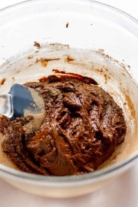 A glass bowl containing thick chocolate batter being mixed with a black spatula.