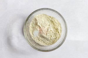 A glass bowl containing a mixture of dry ingredients, including flour and a small amount of pink and white powders, on a white surface.