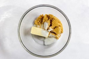A glass bowl containing a stick of butter, brown sugar, and white granulated sugar on a white surface.