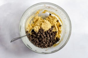 A glass bowl with cookie dough and chocolate chips, with a spoon partially submerged in the mixture, on a white surface.
