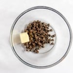 A clear glass bowl containing chocolate chips and a pat of butter on a white surface.