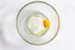 A glass bowl containing an egg yolk, egg white, and a mound of white sugar on a white surface.