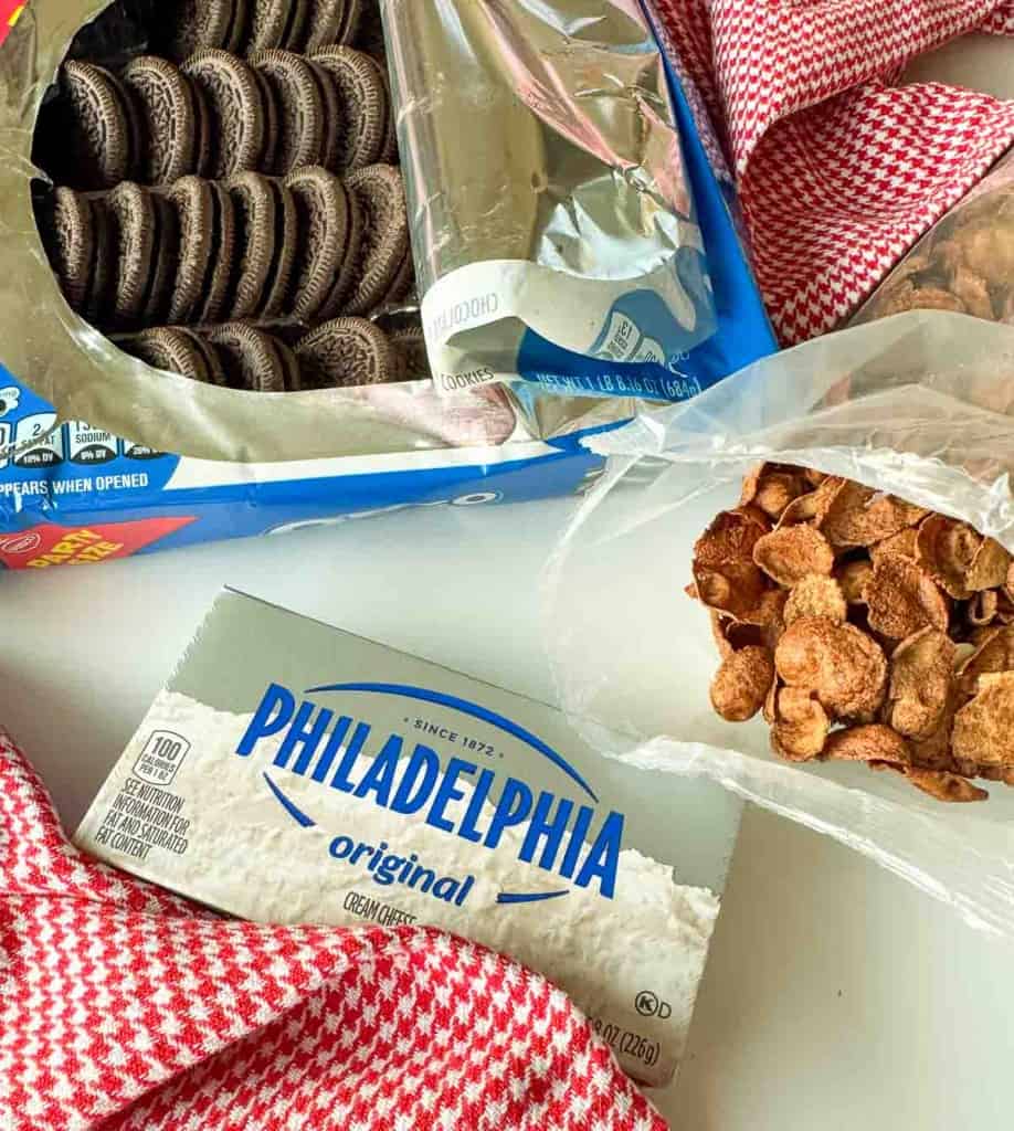An open package of sandwich cookies, a block of Philadelphia cream cheese, and a bag of cornflakes on a red and white towel—perfect ingredients for delicious Chocolate Pinecone Truffles.