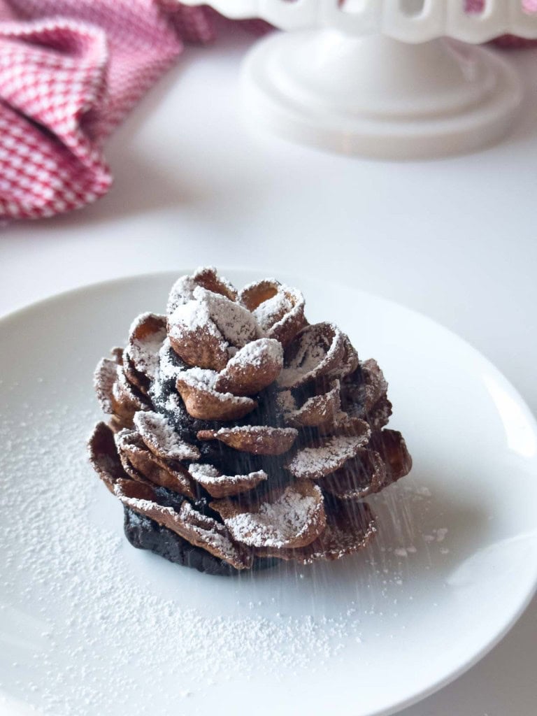 A chocolate pine cone-shaped dessert, known as Chocolate Pinecone Truffles, is dusted with powdered sugar and sits elegantly on a white plate.