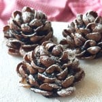 Three Chocolate Pinecone Truffles dusted with powdered sugar are displayed on a white surface, with a pink checkered cloth in the background.