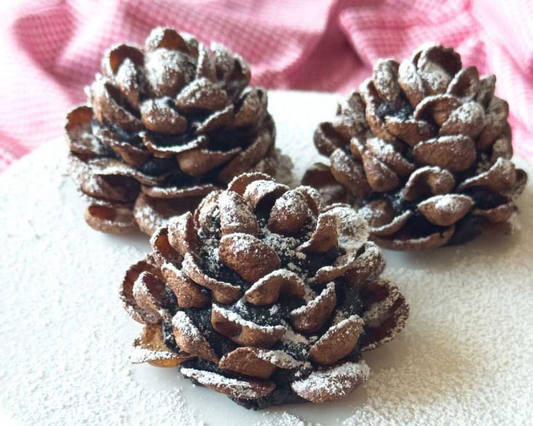 Three Chocolate Pinecone Truffles dusted with powdered sugar are displayed on a white surface, with a pink checkered cloth in the background.