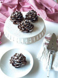 Four Chocolate Pinecone Truffles dusted with powdered sugar are displayed on a white cake stand and plate, with a pie server and a red-checkered cloth nearby.