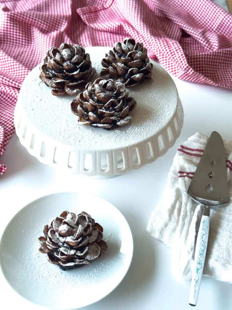 Four Chocolate Pinecone Truffles dusted with powdered sugar are displayed on a white cake stand and plate, with a pie server and a red-checkered cloth nearby.