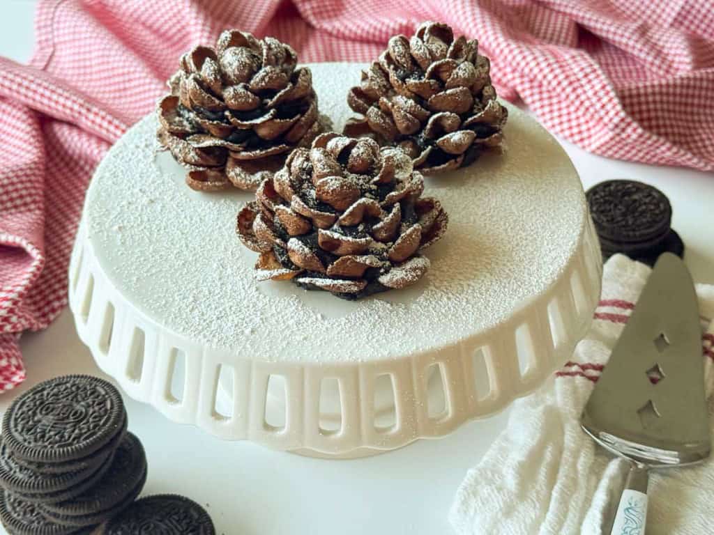Three Chocolate Pinecone Truffles dusted with powdered sugar are displayed on a white cake stand, with Oreos, a cake server, and red checkered cloths nearby.