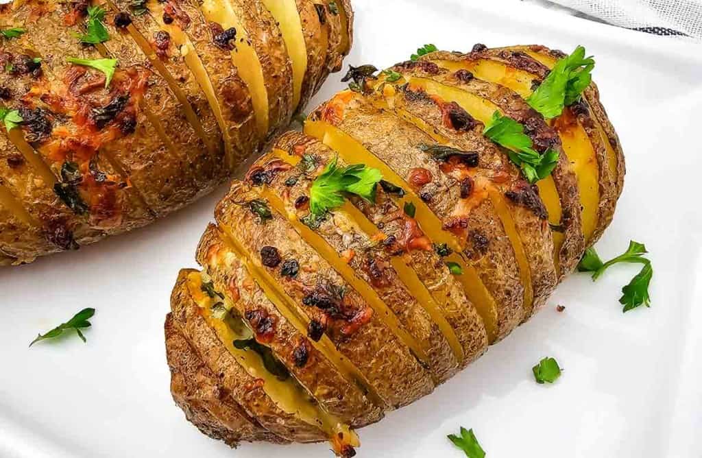 Two Hasselback potatoes topped with melted cheese and herbs on a white plate, garnished with fresh parsley.