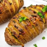 Two Hasselback potatoes topped with melted cheese and herbs on a white plate, garnished with fresh parsley.