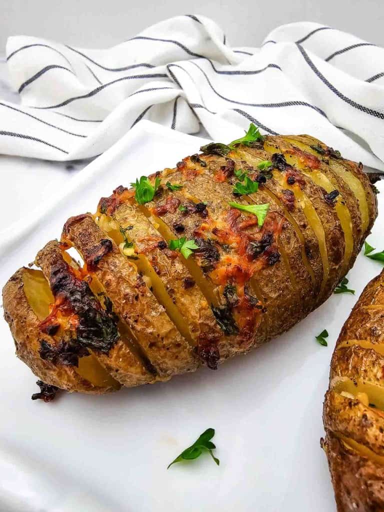 A sliced baked potato with herbs and melted cheese, garnished with chopped parsley, sits on a white surface with a striped cloth in the background.