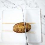 A potato on a white cutting board is being sliced with a knife, guided by two chopsticks placed on either side to prevent cutting all the way through.