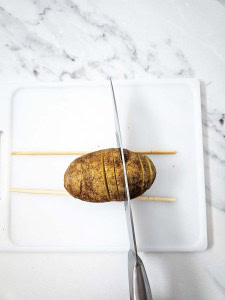 A potato on a white cutting board is being sliced with a knife, guided by two chopsticks placed on either side to prevent cutting all the way through.