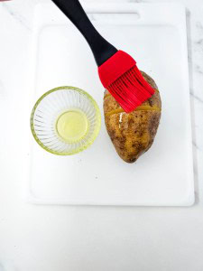 A potato is being brushed with oil using a red silicone brush, next to a small glass bowl of oil, on a white cutting board.