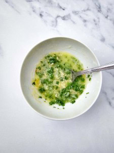 A white bowl containing a mixture of chopped herbs, grated garlic, and oil with a spoon on a marble surface.