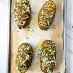 Four sliced Hasselback potatoes topped with melted cheese and herbs on a parchment-lined baking tray.