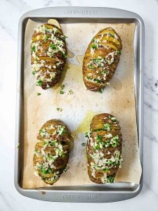 Four sliced Hasselback potatoes topped with melted cheese and herbs on a parchment-lined baking tray.