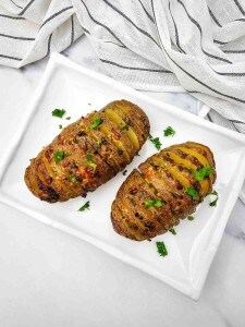 Two Hasselback potatoes garnished with chopped parsley are served on a rectangular white plate with a striped cloth in the background.