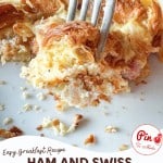 A fork cuts into a ham and swiss croissant breakfast casserole, revealing the flaky layers of croissant, savory ham, and melted Swiss cheese on a plate.