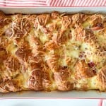 A baked ham and swiss croissant breakfast casserole with melted cheese and visible bits of bacon in a rectangular white dish on a red and white striped cloth.