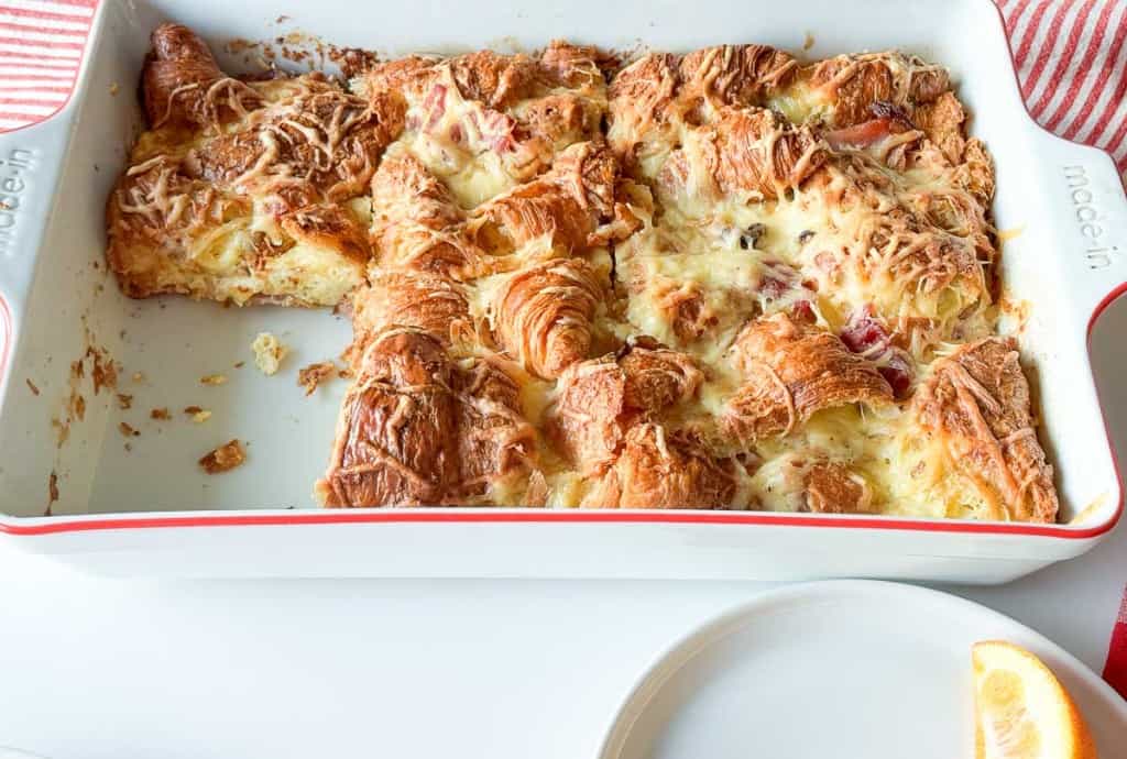 A white baking dish with partially served ham and swiss croissant breakfast casserole topped with melted cheese, resting on a white surface with a red-striped cloth nearby.