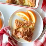 A slice of ham and swiss croissant breakfast casserole and two orange slices on a white plate, next to a red and white checkered napkin.