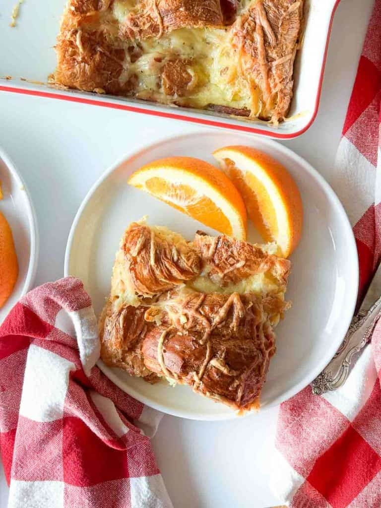 A slice of ham and swiss croissant breakfast casserole and two orange slices on a white plate, next to a red and white checkered napkin.